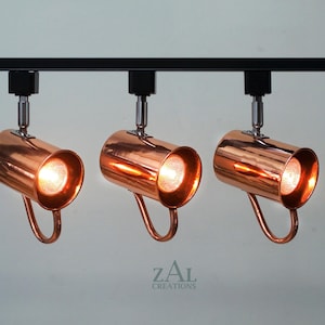 May include: Three copper track lights with black track and white ceiling mount. The lights are on and casting a warm glow.