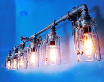 May include: A six-light industrial style wall sconce with clear glass beer mug-shaped light fixtures. The sconce is made of metal pipes and has a rustic look. ZAL Creations is printed on the wall behind the sconce.