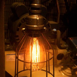 May include: A caged light bulb with a warm, glowing filament hangs from a metal pipe. The light bulb is surrounded by a metal cage, and the pipe is attached to a wall.