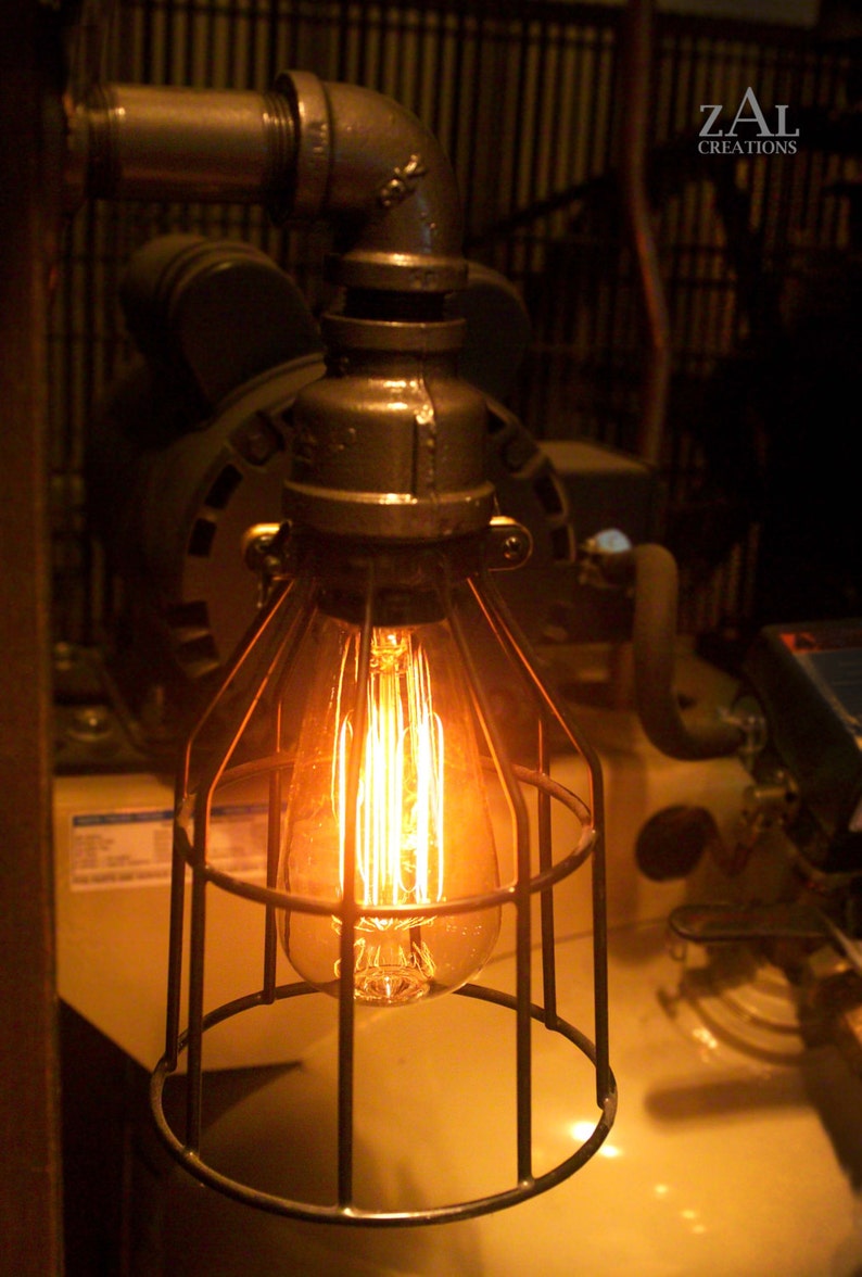 May include: A caged light bulb with a warm, glowing filament hangs from a metal pipe. The light bulb is surrounded by a metal cage, and the pipe is attached to a wall.