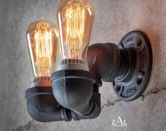 May include: A black metal pipe wall sconce with two Edison bulb light fixtures. The sconce has a black finish and is mounted on a wall. The bulbs are turned on and glowing. The sconce is labelled "ZAL Creations, Chicago, USA."