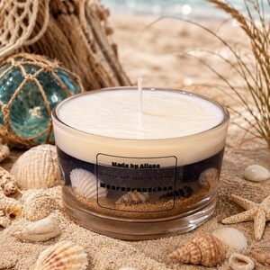 Könnte beinhalten: Ein durchsichtiges Kerzenglas mit weißem Wachs oben und einer dunkelblauen Gelschicht unten, in der Muscheln und Sand eingebettet sind. Das Etikett trägt die Aufschrift Made by Alissa, Gelwax and natural harvest, Meeresrauschen, ESSENTIAL OILS.