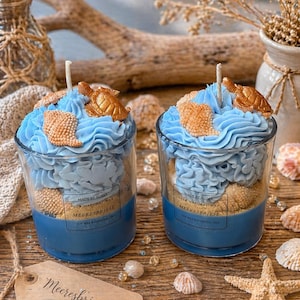 Könnte beinhalten: Zwei Glaskerzen mit blauen, beigen und goldenen Akzenten. Die Kerzen haben eine blaue Basis, eine beige sandartige Schicht und eine blaue, frostingartige Oberseite mit goldenen Schildkröten- und Muscheldekorationen. Das Etikett lautet "Meeresbrise".