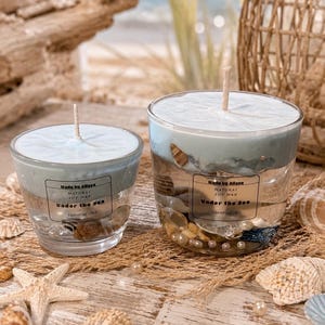Könnte beinhalten: Zwei Kerzen in klaren Gläsern, gefüllt mit hellblauem Sojawachs und eingebetteten Muscheln. Jede Kerze hat ein Etikett mit der Aufschrift Made by Allssa NATURAL SOY WAX Under the Sea ESSENTIAL OILS. Ein einzelner Docht ist mittig platziert.