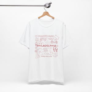 Könnte beinhalten: Ein weißes Kurzarm-T-Shirt mit einem roten Grafikdruck aus Philadelphia-Themen-Strichzeichnungen, darunter eine Brücke, eine Brezel, ein Bus sowie der Text "PENNSYLVANIA", "PHILADELPHIA", "LOVE", "From the 215 with love" und "Philly vibes only".