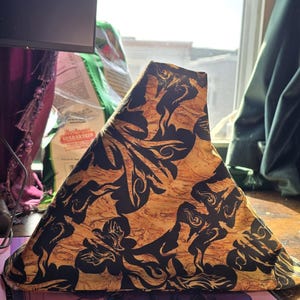 May include: A triangular lampshade with a unique design. The lampshade features a pattern of black swirling shapes against a golden-brown background. The lampshade is sitting on a wooden surface.