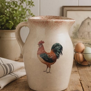May include: A cream-colored ceramic pitcher featuring a detailed illustration of a rooster with red, brown, and dark green feathers. The pitcher has a curved handle, a wide spout, and a speckled reddish-brown finish along the top rim.
