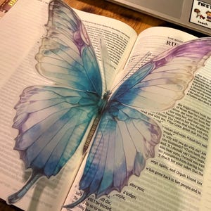 May include: A large, translucent butterfly sticker with blue and purple watercolor-style wings is placed on an open book. The sticker's wings are detailed with veins, and the book's pages are filled with text. A laptop is visible in the background.