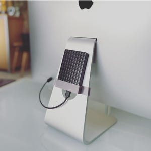 Op de afbeelding: Een zwarte externe harde schijf met een geometrisch patroon is bevestigd aan de achterkant van een zilveren computermonitor. Een zwarte USB-kabel verbindt de harde schijf met de monitor. De monitor heeft een witte achtergrond.