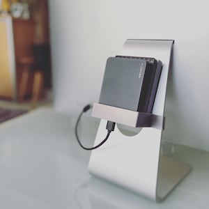 Puede incluir: Un soporte de ordenador plateado que sostiene un disco duro externo Toshiba gris. El disco duro está conectado con un cable USB negro. El soporte está sobre una superficie blanca.