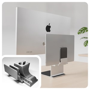 May include: A silver-colored desktop stand designed to hold a tablet and a smartphone. The stand features a dedicated slot for a mobile device, a wider slot for a tablet, and a circular cutout on the top section for a charging cable or accessory.