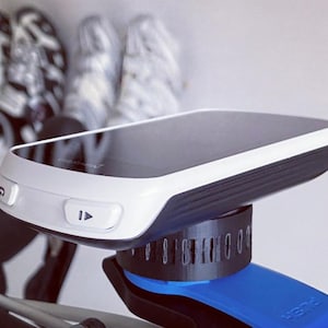 Op de afbeelding: Een zwart-witte fietscomputer met een witte rand en twee knoppen. De computer is gemonteerd op een blauw-zwarte beugel. De achtergrond toont wazige fietsschoenen.