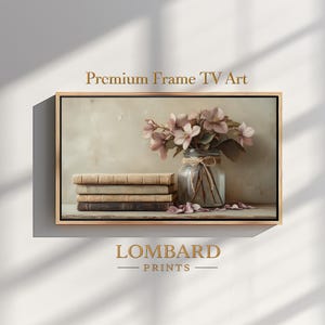 Pode incluir: Impressão emoldurada com uma natureza morta. Apresenta uma pilha de livros antigos, um frasco de vidro com flores cor-de-rosa e pétalas espalhadas. A moldura inclui o texto "Premium Frame TV Art" e "LOMBARD PRINTS".