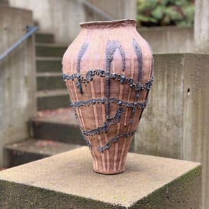 May include: A large, handcrafted ceramic vase with a textured, reddish-brown surface. Vertical lines and dark gray accents adorn the vase, with a decorative chain-like pattern around the middle. The vase is displayed on a concrete pedestal.