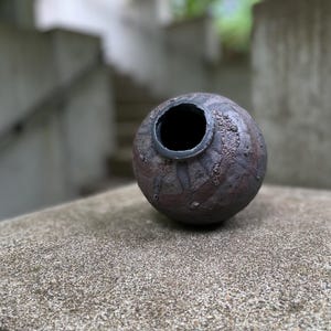 Puede incluir: Un jarrón esférico de cerámica hecho a mano con una superficie oscura y texturizada. El jarrón tiene una boca ancha y abierta con un borde oscuro. El exterior presenta un patrón moteado de tonos marrones y negros, lo que sugiere una estética rústica. Es un objeto decorativo.