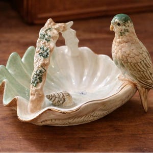 May include: A ceramic seashell-shaped dish with a handle and a perched bird figurine. The dish is white with a gold trim and a light green outer edge. The bird is beige and green.