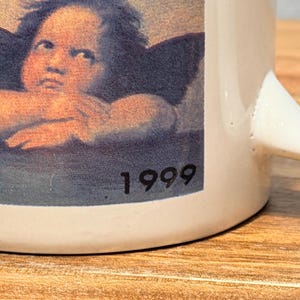 Op de afbeelding: Een witte keramische mok met een afbeelding van een cherubijn met donkere vleugels die zijn kin op zijn handen laat rusten. De print bevat het jaartal 1999 in zwarte tekst in de rechterbenedenhoek van de afbeelding. De mok heeft een gebogen handvat.