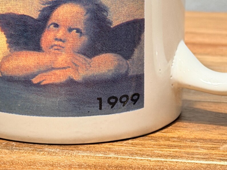 Op de afbeelding: Een witte keramische mok met een afbeelding van een cherubijn met donkere vleugels die zijn kin op zijn handen laat rusten. De print bevat het jaartal 1999 in zwarte tekst in de rechterbenedenhoek van de afbeelding. De mok heeft een gebogen handvat.
