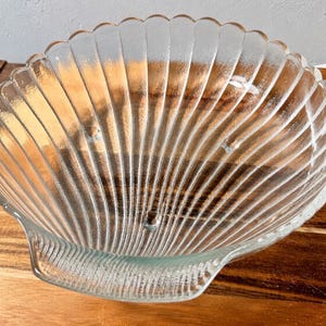 Könnte beinhalten: Eine durchsichtige Glasschale in Muschelform mit gewelltem Rand und geripptem Innenraum. Die Schale hat eine griffartige Verlängerung auf einer Seite. Die Schale steht auf einer Holzoberfläche.