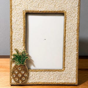 May include: A rectangular picture frame featuring a white beaded surface with gold beaded trim. A decorative pineapple applique is attached to the lower left corner, crafted with green glass beads for the leaves and gold beads with clear crystals.