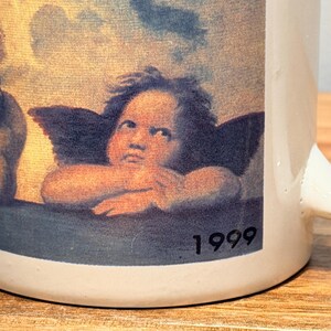 Op de afbeelding: Een witte keramische mok met een gedrukte reproductie van twee cherubijntjes uit de Sixtijnse Madonna van Rafa&euml;l. De afbeelding toont de twee gevleugelde figuren die hun kin op hun handen laten rusten tegen een bewolkte lucht, met het jaartal 1999 in zwart.