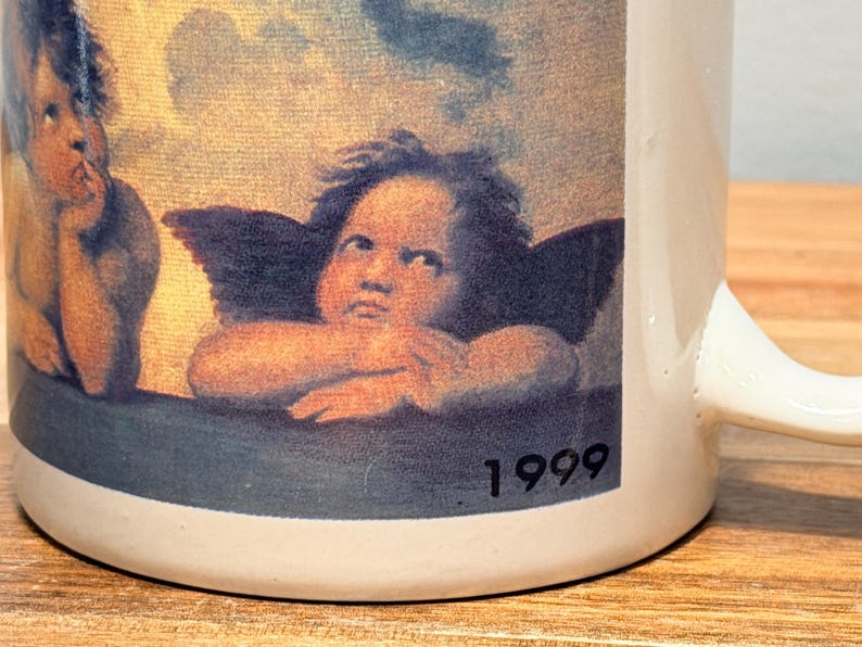 Op de afbeelding: Een witte keramische mok met een gedrukte reproductie van twee cherubijntjes uit de Sixtijnse Madonna van Rafa&euml;l. De afbeelding toont de twee gevleugelde figuren die hun kin op hun handen laten rusten tegen een bewolkte lucht, met het jaartal 1999 in zwart.
