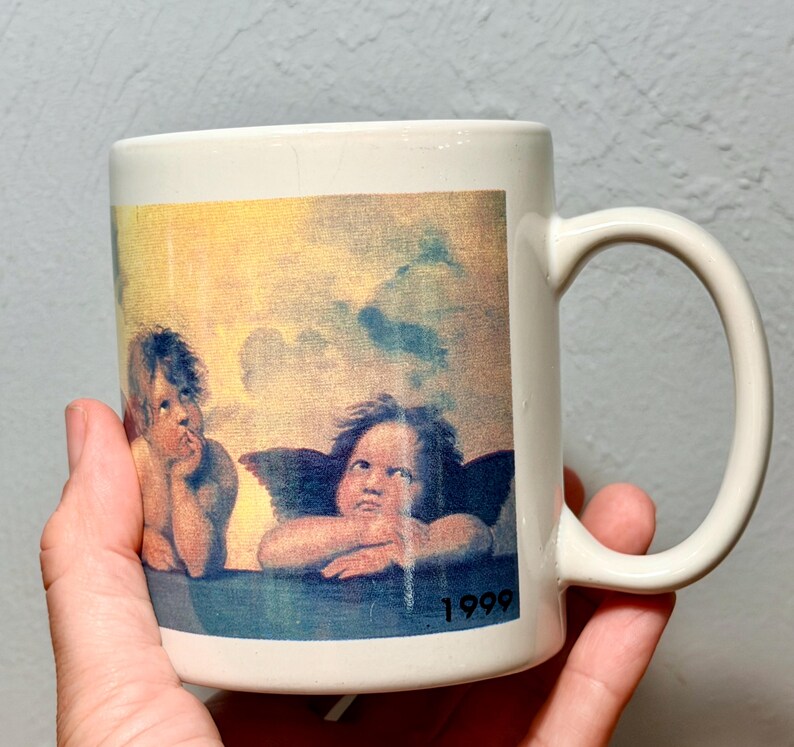 Op de afbeelding: Een witte keramische mok met een gedrukte reproductie van twee cherubijntjes uit de Sixtijnse Madonna van Rafa&euml;l. De afbeelding toont de twee gevleugelde figuren die hun kin op hun handen laten rusten tegen een gele en blauwe lucht, met het jaartal 1999.