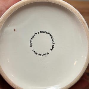 Op de afbeelding: De ronde witte onderkant van een keramische mok met zwarte tekst die DISHWASHER & MICROWAVEABLE SAFE en MADE IN CHINA vermeldt. De onderzijde heeft een gladde, ongeglazuurde beige rand en een klein bruin vlekje op het oppervlak.