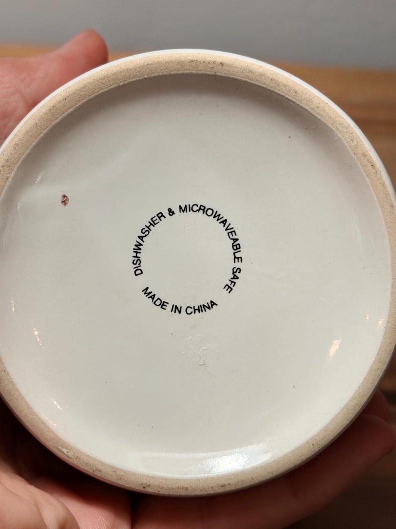 Op de afbeelding: De ronde witte onderkant van een keramische mok met zwarte tekst die DISHWASHER & MICROWAVEABLE SAFE en MADE IN CHINA vermeldt. De onderzijde heeft een gladde, ongeglazuurde beige rand en een klein bruin vlekje op het oppervlak.