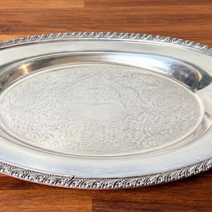Puede incluir: Una bandeja de servicio ovalada de color plateado que presenta un centro liso y pulido con un intrincado patrón floral grabado y un borde decorativo estilo cuerda a lo largo del borde exterior. La bandeja tiene un acabado metálico brillante.