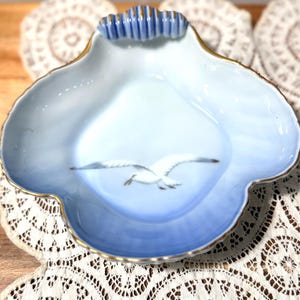 May include: A decorative, light blue ceramic dish with a gold trim and a scalloped edge. The dish features a painted seagull in flight. The handle is a series of vertical blue ridges with gold accents. A charming piece for display.