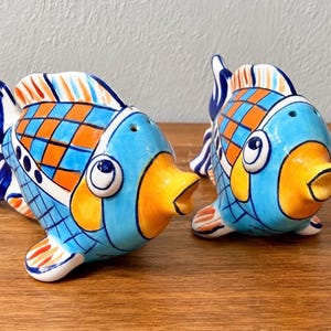 May include: A pair of ceramic fish-shaped salt and pepper shakers featuring a vibrant hand-painted pattern of blue and orange checkered scales, yellow faces, and dark blue fins. Each fish has small holes on the top for dispensing seasoning.
