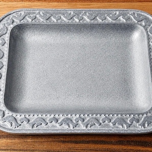 May include: A rectangular, silver-colored tray with a decorative border featuring a repeating pattern of stylized shapes. The tray is set on a wooden surface, showcasing its design and texture.