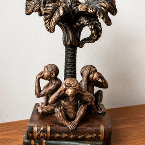 May include: A bronze-toned sculpture featuring three monkeys sitting on a stack of two books. One monkey covers its ears, one covers its eyes, and one covers its mouth. A palm tree structure rises from the center of the monkeys, with gold highlights.