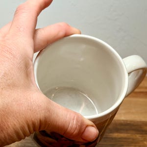 Op de afbeelding: Een witte keramische mok met een gladde, glanzende binnenkant en een gebogen handvat aan de zijkant. De mok heeft een cilindrische vorm met een brede opening aan de bovenkant, wat een schone, lege kom laat zien. De buitenkant is onderaan zichtbaar.