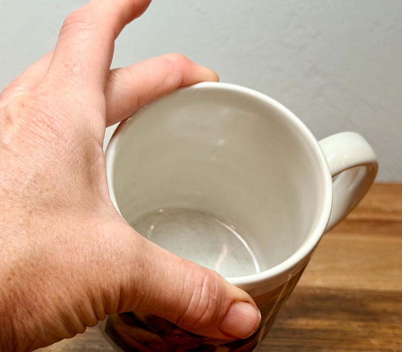 Op de afbeelding: Een witte keramische mok met een gladde, glanzende binnenkant en een gebogen handvat aan de zijkant. De mok heeft een cilindrische vorm met een brede opening aan de bovenkant, wat een schone, lege kom laat zien. De buitenkant is onderaan zichtbaar.