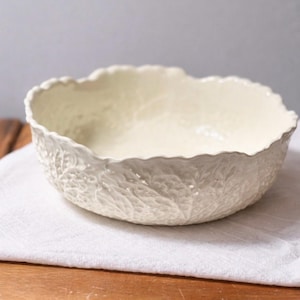 May include: Off-white ceramic bowl with a textured, leaf-like pattern. The bowl has a scalloped rim and sits on a white cloth over a wooden surface. The bowl's interior is smooth and light-colored.