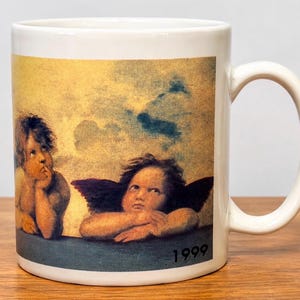 Op de afbeelding: Een witte keramische mok met een gedrukte reproductie van twee cherubijntjes uit de Sixtijnse Madonna van Rafa&euml;l. De afbeelding toont twee gevleugelde figuren die hun kin op hun handen laten rusten tegen een gele luchtachtergrond, met het jaartal 1999 in het zwart gedrukt.