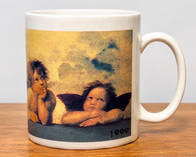 Op de afbeelding: Een witte keramische mok met een gedrukte reproductie van twee cherubijntjes uit de Sixtijnse Madonna van Rafa&euml;l. De afbeelding toont twee gevleugelde figuren die hun kin op hun handen laten rusten tegen een gele luchtachtergrond, met het jaartal 1999 in het zwart gedrukt.