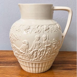May include: A cream-colored ceramic pitcher featuring a raised relief design of two figures with a dog in a landscape setting. The upper portion displays a grape vine motif, while the base is decorated with vertical fluted lines and a handle.