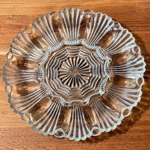 May include: A clear glass serving plate featuring a central circular section with a radiating sunburst pattern, surrounded by a wider outer rim decorated with a series of elongated, fan-shaped fluted panels and a scalloped, beaded edge.