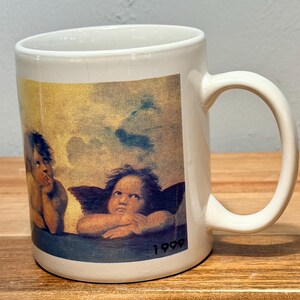 Op de afbeelding: Een witte keramische mok met een gedrukte reproductie van de cherubijnen van Rafa&euml;ls Sixtijnse Madonna. De afbeelding toont twee gevleugelde figuren tegen een bewolkte gele en blauwe lucht, met het jaartal 1999 in zwart rechtsonder gedrukt.