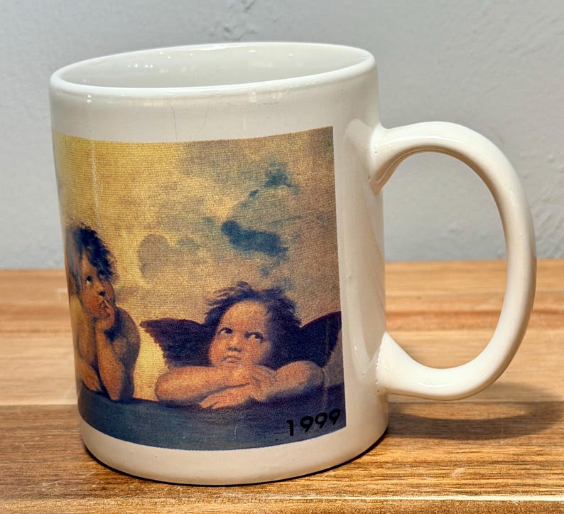 Op de afbeelding: Een witte keramische mok met een gedrukte reproductie van de cherubijnen van Rafa&euml;ls Sixtijnse Madonna. De afbeelding toont twee gevleugelde figuren tegen een bewolkte gele en blauwe lucht, met het jaartal 1999 in zwart rechtsonder gedrukt.