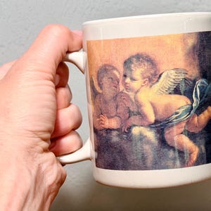 Op de afbeelding: Een witte keramische mok met een gedrukte reproductie van een klassiek schilderij met twee gevleugelde cherubijntjes. E&eacute;n cherubijntje zit terwijl de andere voorover leunt, gedrapeerd in een blauwe doek tegen een warme, goudkleurige achtergrond.
