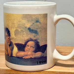 Op de afbeelding: Een witte keramische mok met een gedrukte reproductie van de twee cherubijntjes uit de Sixtijnse Madonna van Rafa&euml;l. De afbeelding toont twee gevleugelde figuren die hun kin op hun handen laten rusten tegen een bewolkte lucht, met het jaartal 1999.