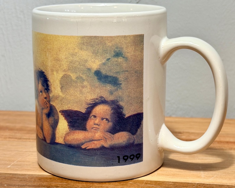 Op de afbeelding: Een witte keramische mok met een gedrukte reproductie van de twee cherubijntjes uit de Sixtijnse Madonna van Rafa&euml;l. De afbeelding toont twee gevleugelde figuren die hun kin op hun handen laten rusten tegen een bewolkte lucht, met het jaartal 1999.