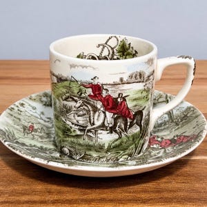 May include: A ceramic teacup and saucer set featuring a detailed transferware print of a fox hunt scene. The design depicts riders in red coats on horseback across a green landscape. The interior of the cup shows a decorative letter B motif.