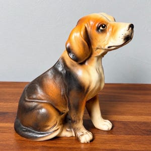 May include: A ceramic figurine of a beagle dog in a seated position. The dog features a tricolor coat with patches of black, tan, and white. It has long, drooping ears, dark eyes, and a focused expression, with its head turned to the right side.