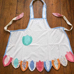 May include: White apron with blue trim and a decorative floral design. The apron features a heart-shaped patch and a row of colorful, petal-shaped appliqués along the bottom edge. The ties are cream-colored with red and white striped accents.