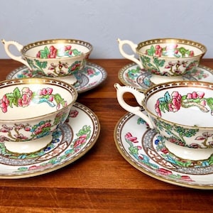 May include: A set of four porcelain teacups and matching saucers featuring a cream-colored base with hand-painted pink floral motifs, green foliage, and intricate brown geometric border patterns, all finished with gold-toned rims.